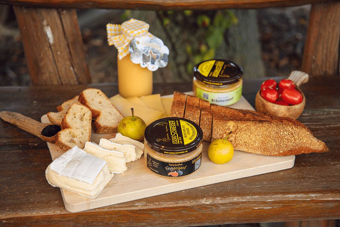Auf einem Holztisch liegt eine Brettljause mit Feigensenf, Brot, Käse, Tomaten und mehr.