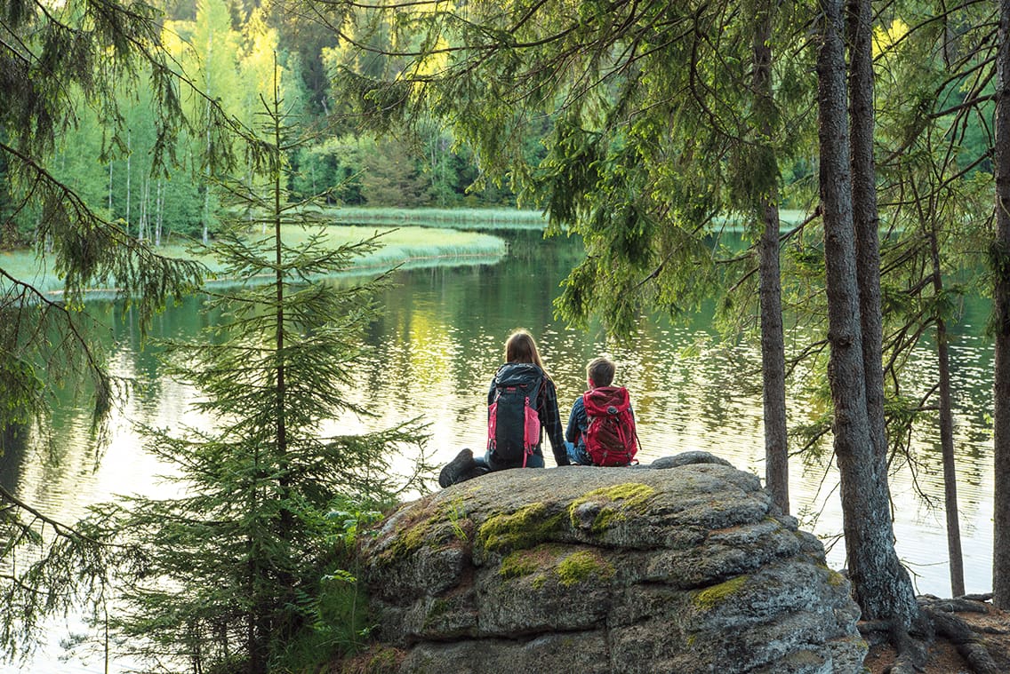 Kinder sehen sich auf den Stein sitzend im Wald einen See an.