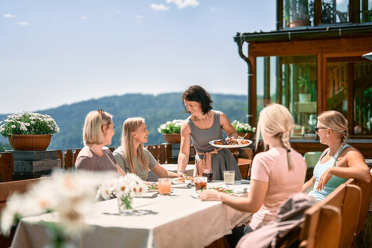 Eine Kellnerin serviert vier Damen frische steirische Gerichte auf der Alm in Sommer.