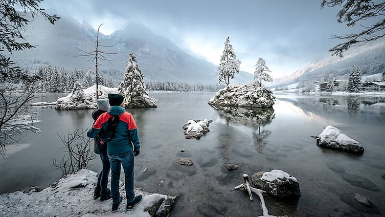 Winterlandschaft im Bergsteigerdorf Ramsau in Bayern – zwei Personen blicken auf verschneite Bäume und Felsen im zugefrorenen Hintersee, umgeben von schneebedeckten Bergen.