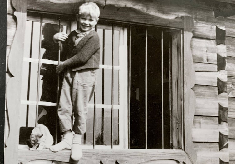 Hansi Hinterseer mit einer Katze als Kind. Er steht auf einem Fensterrahmen eines Holzhauses.