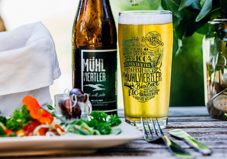 Eine Flasche Bier und ein gefülltes Glas Bier stehen auf einem Tisch, unscharf sieht man einen Teller mit Salat.