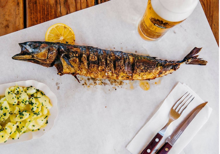 ein köstlicher Steckerfisch liegt auf einem Stück Backpapier, daneben steht ein Erdäpfelsalat mit Schnittlauch und ein großes Glas Bier.
