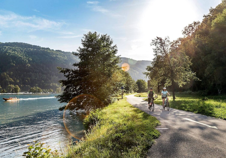 Zwei Radfahrer im Sommer fahren auf einem Radweg entlang des Wasser, der Donau, die Sonne scheint, alles ist grün.