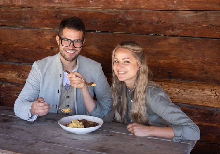 Daniele Zingerele und Paula Bründel essen gemeinsam aus einem Teller ein Rindergulasch mit Polenta und sitzen dabei vor einer Holzhütte auf einer Holzbank mit Holztisch.