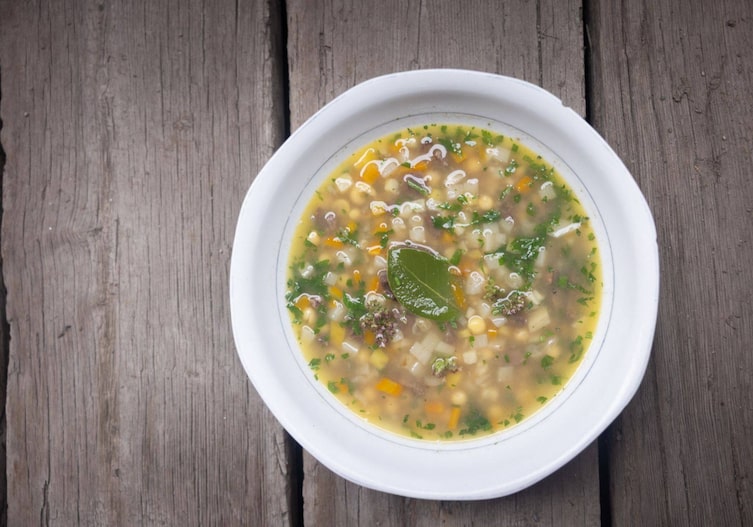 rollgerstesuppe mit Wurzelgemüse (gewürfelt) mit frischen Kräutern und einem Lorbeerblatt. Die Suppe ist in einem tiefen Teller angerichtet und steht auf einer Holzunterlage.
