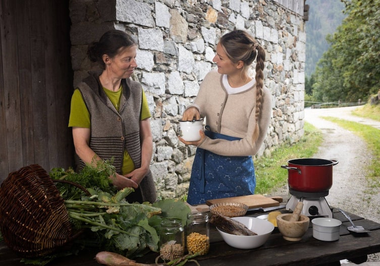 Sabine Prenn und Paula Bründl stehen mit einem Holztisch auf dem alle Zutaten für eine Graupensuppe aufgereiht sind, in der hofeinfahrt. Sie haben auch einen Kocher und einen Topf zum Kochen dabei.