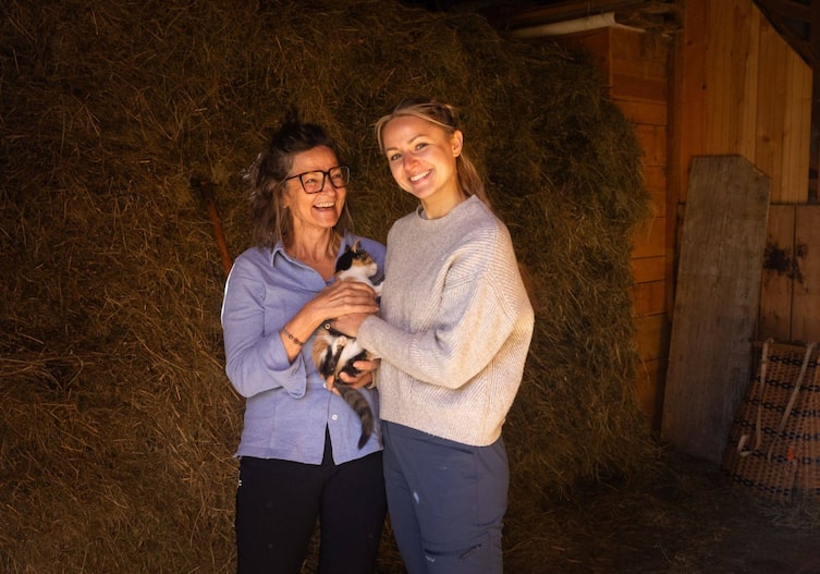 Heustall, zwei Frauen, die in die Kamera lachen und ein kleines Katzerl streicheln.