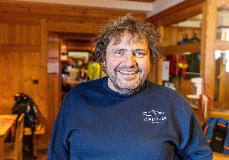 Siegfried Karl lacht in die Kamera und steht in einer Stube in einer Hütte