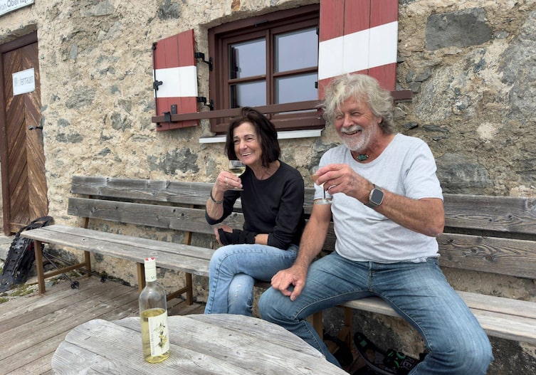 Wirtsleut Studer vor der Hütte auf einer Holzbank. Sie trinken ein Glaserl Weißwein und lachen dabei.