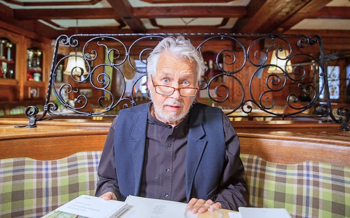 Hans Krumpmüller sitzt in einer Gaststube, hat eine Lesebrille auf der Nase sitzen und aufgeschlagene Bücher vor sich liegen.