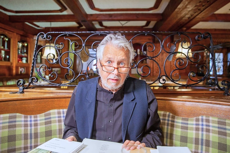 Hans Krumpmüller sitzt in einer Gaststube, hat eine Lesebrille auf der Nase sitzen und aufgeschlagene Bücher vor sich liegen.