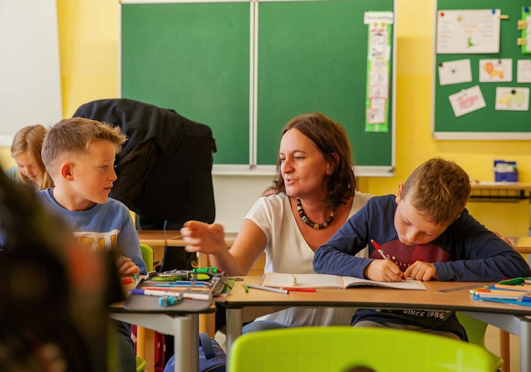 Lehrerin und Kinder in einem Klassenzimmer, die Lehrerin erklärt etwas, sitzt zwischen den Kindern am Tisch, ein Bub schreibt etwas in ein Heft, der andere hört zu.