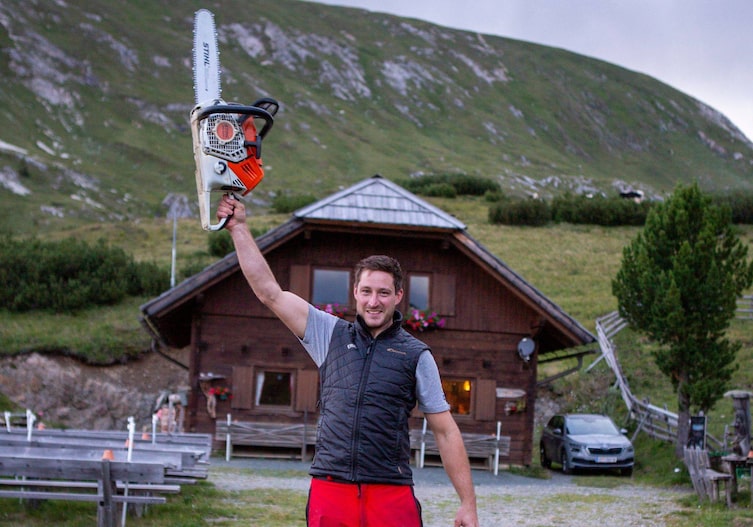 Jürgen Erlacher streckt eine Stihl Motorsäge mit dem Arm gerade in Luft und lächelt dabei. Er steht auf der Alm vor der Erlacher Bockhütte. Es ist Sommer.