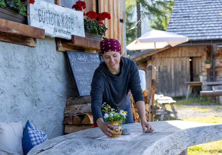Eine Frau mit einem Kopftuch stellt auf einen Holztisch vor einer Almhütte eine kleine Vase mit frischen Almwiesenblumen und lächelt dabei.