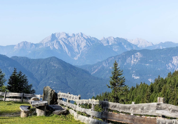 Auf einer Anhöhe auf der Alm, eine Wiese mit einem alten Holzzaun begrenzt mit einem Tisch und Sitzbänken aus Holz. Man hat einen Rundumblick ins Tal und am anderen Ende Hohe Berge zum Teil mit Wald. Es ist Sommer.
