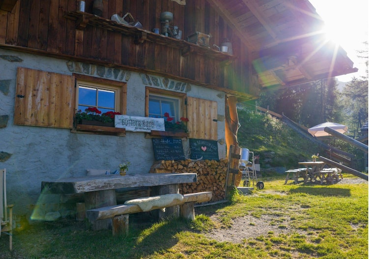 Eine Almhütte mit viel Holz, einer Sitzgelegenheit vor der Tür, ein Stapel Brennholz an der Wand. Es ist Sommer.