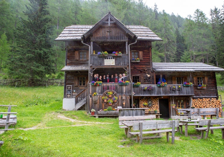 Eine sehr alte Almhütte mit zwei Stockwerken und Blumenkisteln, auf der Veranda im ersten Stock steht ein Chor. Es ist Sommer.