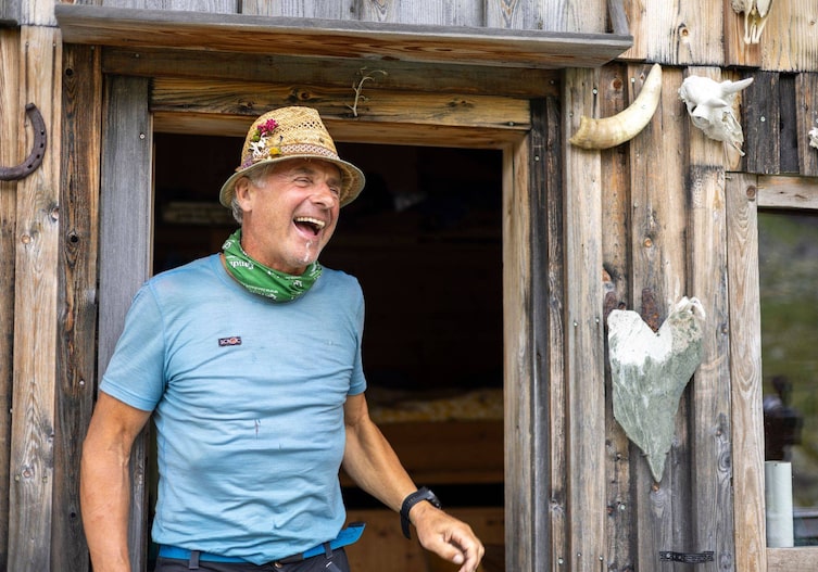 Ein älterer Mann mit grau-weißem Haar und Sonnenhut steht lachend beim Eingang einer Almhütte.
