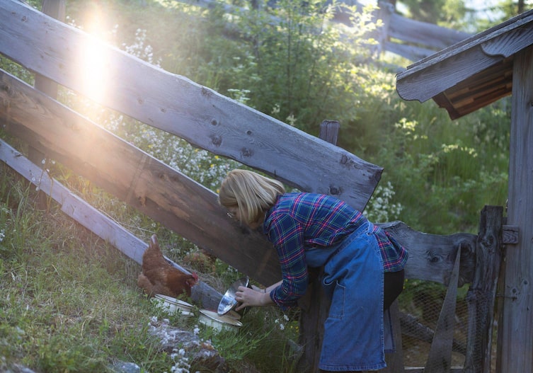 Eine blonde Frau mit Brille und halblangen Haaren und einer Jeansschürze gibt einem Huhn, das in der Wiese stitzt etwas zu essen.