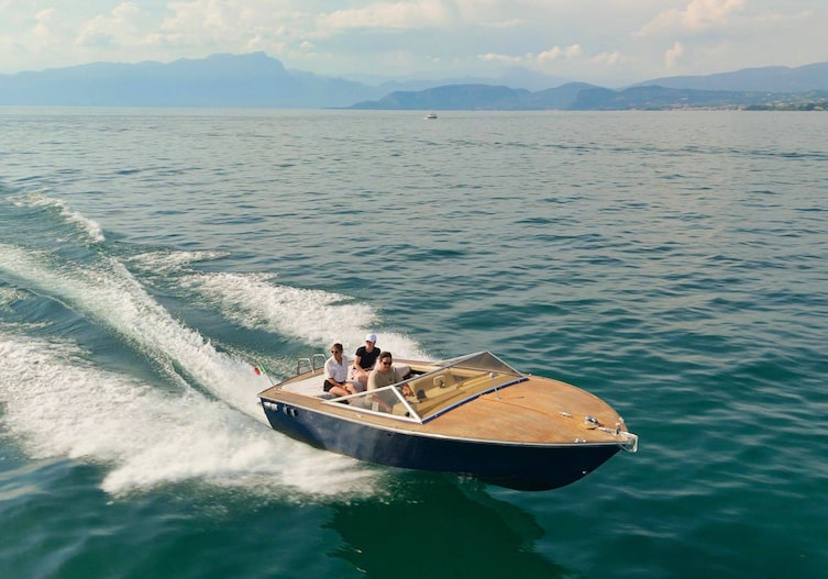 Ein kleines blaues Rennboot fährt auf dem Gardasee. Das Boot wird von einem Mann gelenkt und zwei Frauen sitzen hinter ihm im Boot. Es ist Sommer.