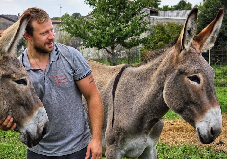 Der Eselzüchter Luca steht auf einer Wiese mit zwei Eseln, einen davon hält er am Kopf