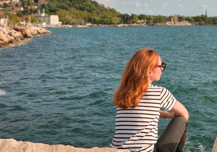 Eine Frau mit langem roten Haar sitzt mit dem Rücken zur Kamera am Meer