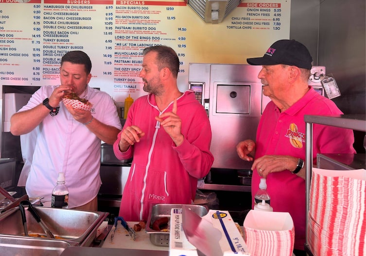 Fritz und Tamino Grampelhuber mit Pinks Hot Dogs Chef und Besitzer Richard Pink im Foodtruck, Tamino verkostete ein Hot Dog und die anderen beiden schauen ihm aufmerksam dabei zu.