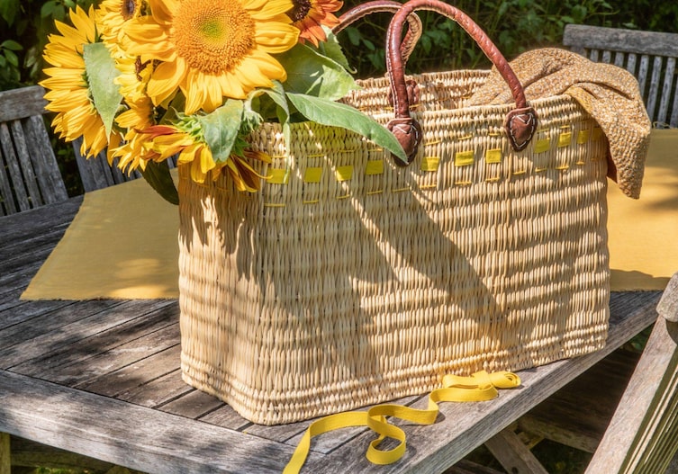 Jutekorb mit gelben Bändern verziert. Im Korb stecken Sonnenblumen. Alles zusammen steht auf einer Gartengarnitur aus Holz.