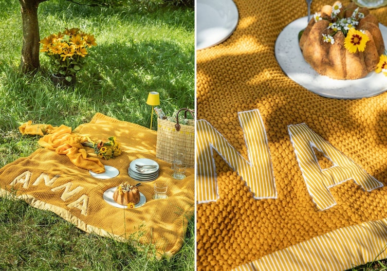 Eine gelbe Picknickdecke, die mitten in der Wiese liegt, mit Tellnern, einem Gugelhupf, der Jutekorb und gelben Blumenstrauß. Alles liegt unter einem Baum.