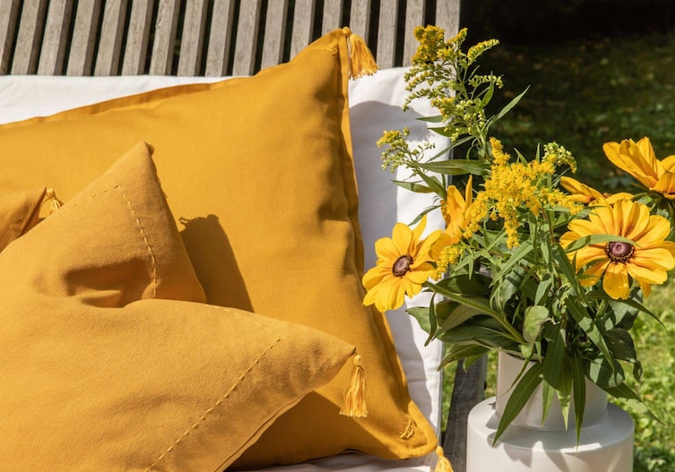 Gelbe Zierpölster mit Quasten verziert. Die Pölster liegen auf einer Gartengarnitur. Daneben steht eine Keramikvase mit gelben Blumen.
