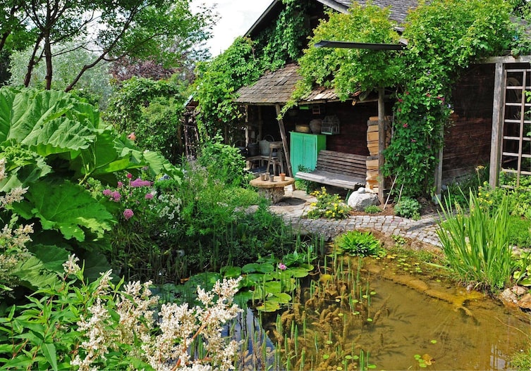 Kleiner Gartenteich mit viel Bewuchs rundherum, im Hochsommer, dahinter steht eine kleine sehr alte Holzhütte mit einer Holzbank