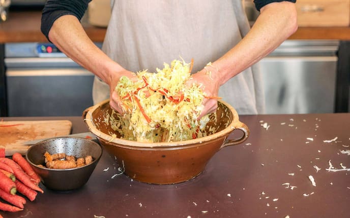 Frau mit aufgestrickten Ärmen knetet in einem braunen Tohntopf Sauerkraut. Daneben steht eine Schüssel mit Ingwerwurzeln und rote Karotten.
