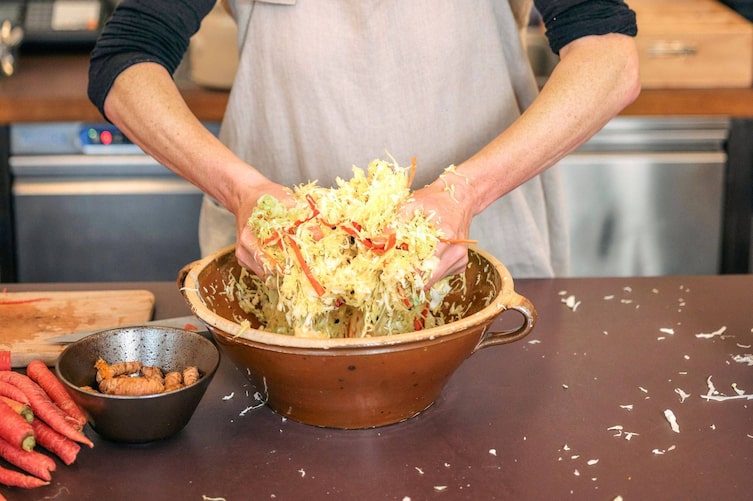 Frau mit aufgestrickten Ärmen knetet in einem braunen Tohntopf Sauerkraut. Daneben steht eine Schüssel mit Ingwerwurzeln und rote Karotten.