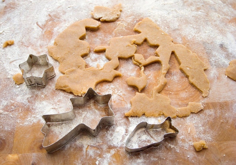 Keksteig auf einer bemehlten Holzfläche, Kekse sind ausgestochen, Ausstecker in Form von Sternen und ein Fisch liegen daneben.