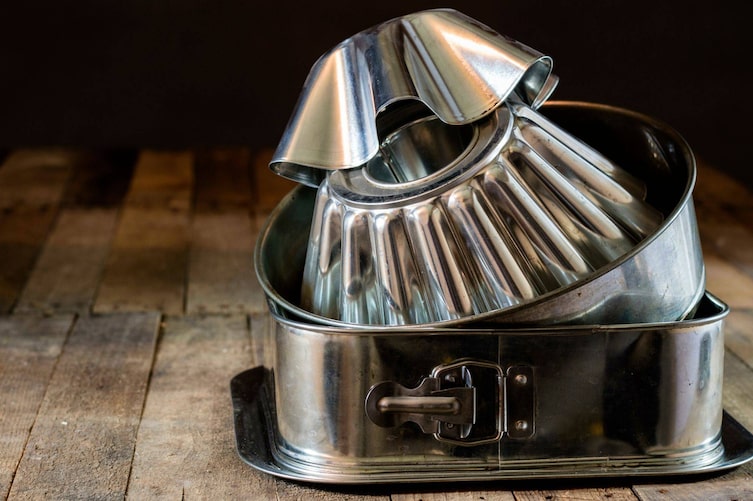 Gewellte Kuchenformen, Tortenspringform rund und eckig, Serviertablett, alles aus Blech, steht auf einem Holzuntergrund und ist übereinander gestapelt.