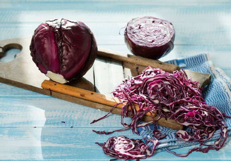 ein ganzer Rotkohl, ein halber Rotkohl und gehobeltes Rotkraut liegen auf einem türkisen Holzuntergrund mit Krauthobel und einem blauen Geschirrtuch