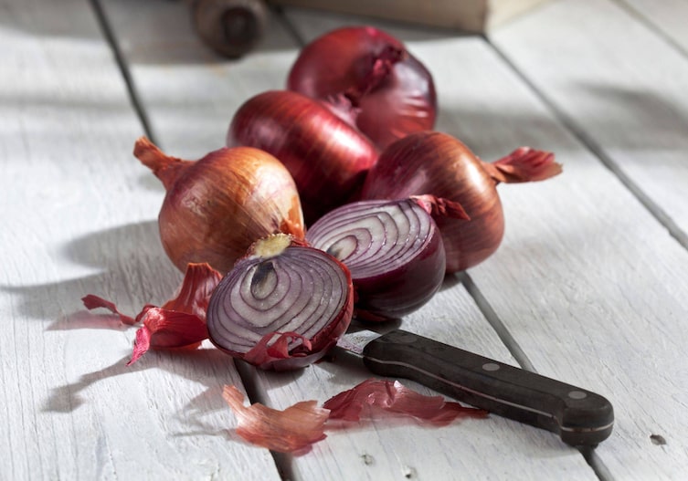 Rote Zwiebel auf einem weißen Holzuntergrund. Eine Zwiebe ist in der Mitte aufgeschnitten. Ein Messer liegt unter der aufgeschnittenen Zwiebel.