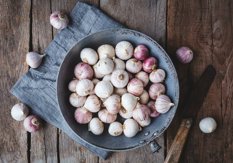 Chinesischer Knoblauch in einer alten Pfanne ohne Stiel, steht auf einer Holzunterlage, daneben liegt ein altes Messer und die Pfanne steht auf einer blauen Stoffserviette.