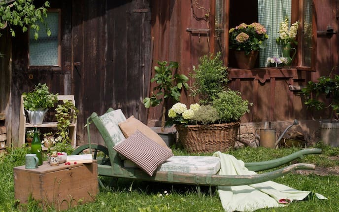 Grüne Einradschubkarre aus Holz, mit bezogenem Schaumstoff und Pölstern. Als Beistelltisch dient eine alte Holzkiste. Alles steht in der Wiese, es ist Sommer. Dahinter ist eine alte braune Gartenhütte mit Fenster zu sehen. Davor stehen Blumen und Kräuter in Töpfen und Körben.