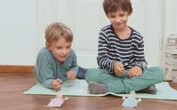 Zwei Buben liegen am Boden auf einem Teppich und spielen ein Schildkrötenrennen, mit selbst gebastelten Schildkröten aus Papier an einer Schnur.