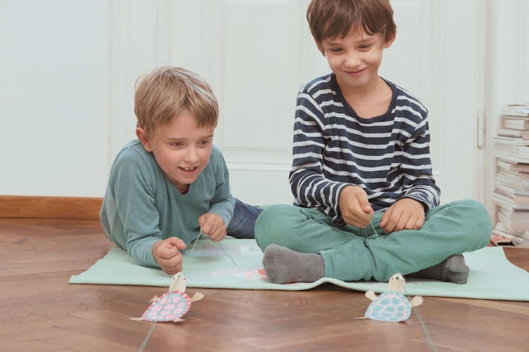 Zwei Buben liegen am Boden auf einem Teppich und spielen ein Schildkrötenrennen, mit selbst gebastelten Schildkröten aus Papier an einer Schnur.