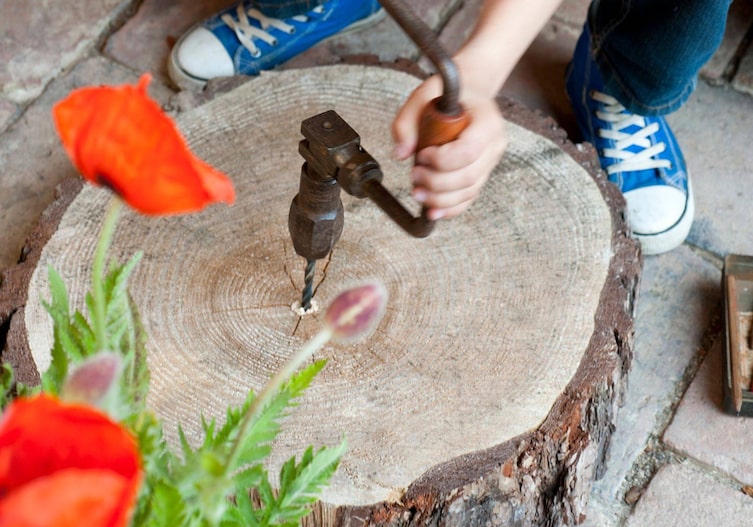 Ein Kind das mit einem Handbohrer ein Loch in die Mitte einer Holzscheibe bohrt. Die Scheibe liegt am Boden.
