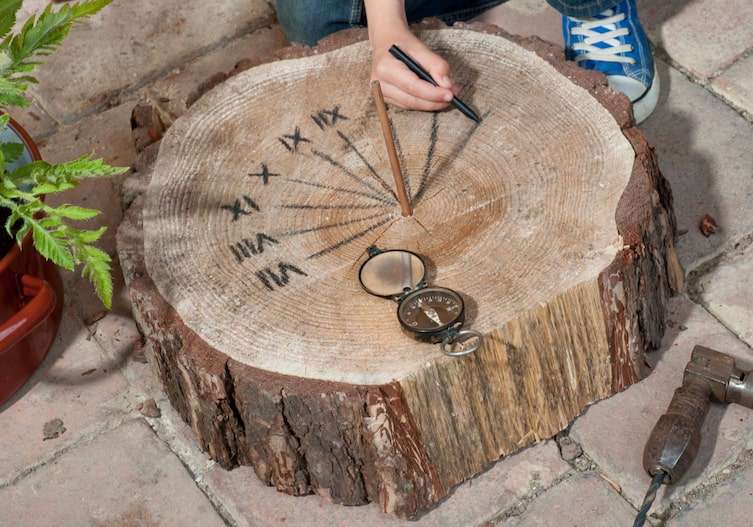 Eine Kinderhand, die auf der Holzscheibe, in der in der Mitte ein Bleistift steckt, die Schatten des Bleistifts, Stunde für Stunde markiert. Daneben liegt ein Kompass.