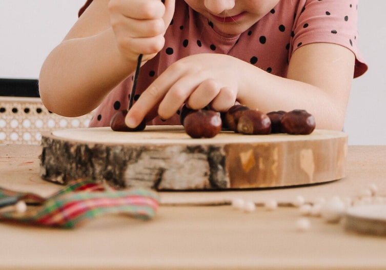 Ein Kind, das mit dem Kastanienbohrer von oben in eine Kastanie drückt. Die Kastanie liegt auf einer Holzscheibe.