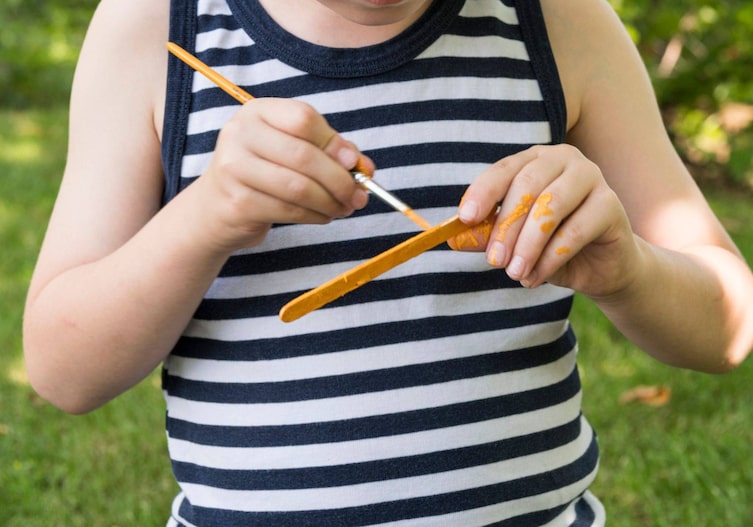 Kinderhände, die mit einem Haarpinsel gelbe Farbe auf ein Eisstäbchen auftragen.