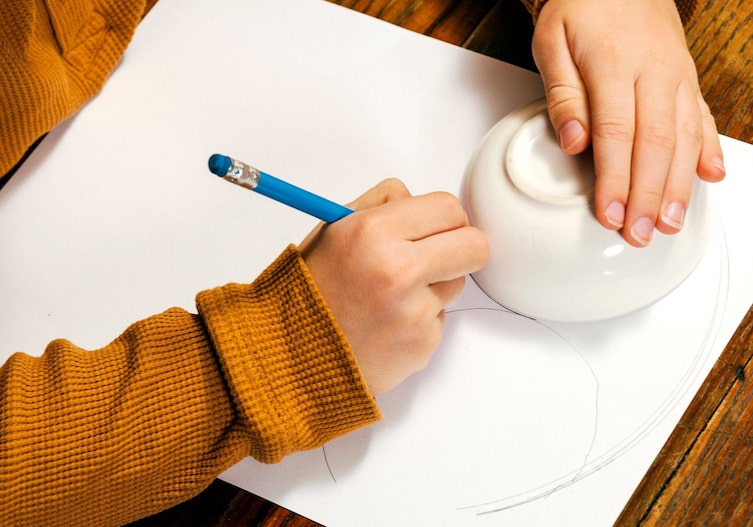 Kinderhände, die die Grundform der Fuchsmaske mit einem Schüsserl als Vorlage auf ein Stück Papier zeichnet