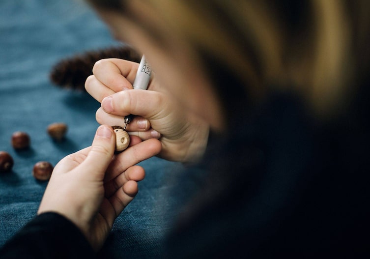 Kinderhände, die eine kleine Holzkugelhalten und mit einem Fineliner Augen auf die Kugel malen.