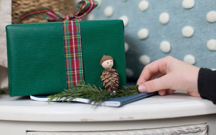 Wichtel aus Bockerl, Holzkugel als Gesicht und Eichelkappe als Hut - auf einem Tannenzweig vor einem Geschenk aufgestellt.