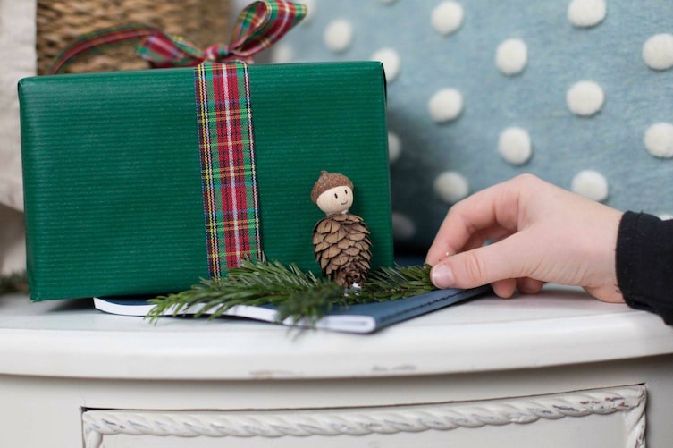 Wichtel aus Bockerl, Holzkugel als Gesicht und Eichelkappe als Hut - auf einem Tannenzweig vor einem Geschenk aufgestellt.